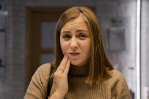 Une femme caucasienne exprimant une gêne due à un mal de dents, portant un pull marron à l'intérieur.