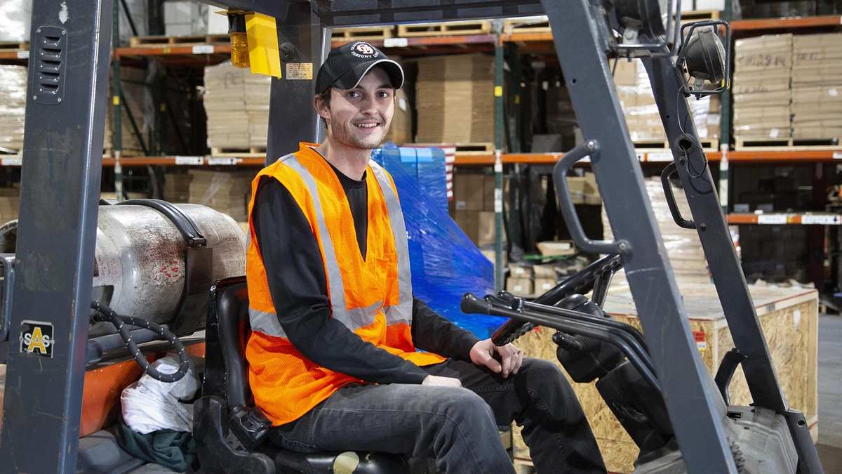 Homme souriant sur chariot élévateur dans entrepôt, vêtu d'un gilet de sécurité