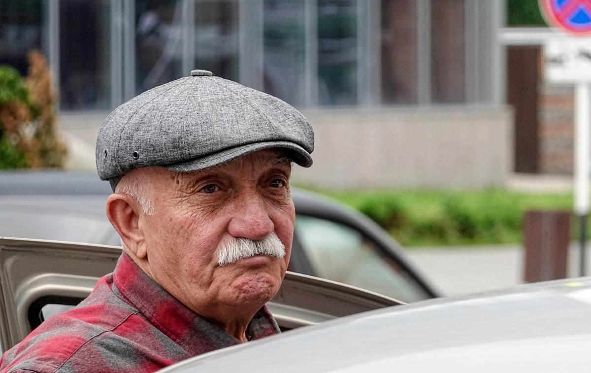 Homme âgé avec moustache et casquette grise, assis dans une voiture