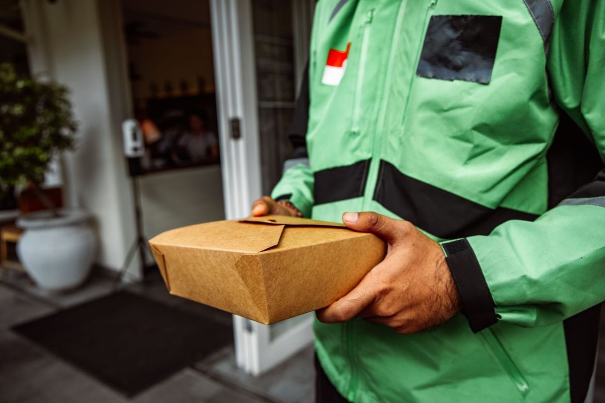 Livreur en uniforme vert noir portant un badge rouge blanc noir, tenant une boîte en carton devant une maison