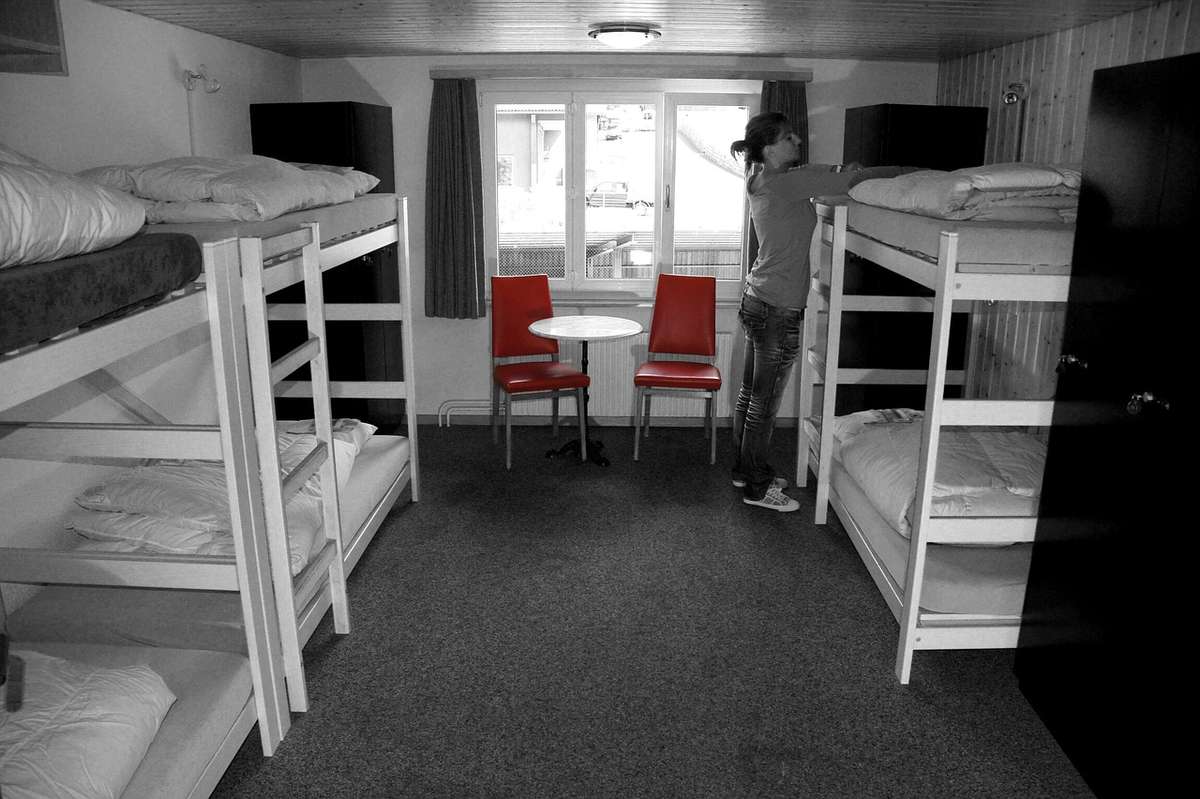 Une chambre avec lits superposés, table, chaises et une personne devant la fenêtre Une chambre avec lits superposés, table, chaises et une personne devant la fenêtre